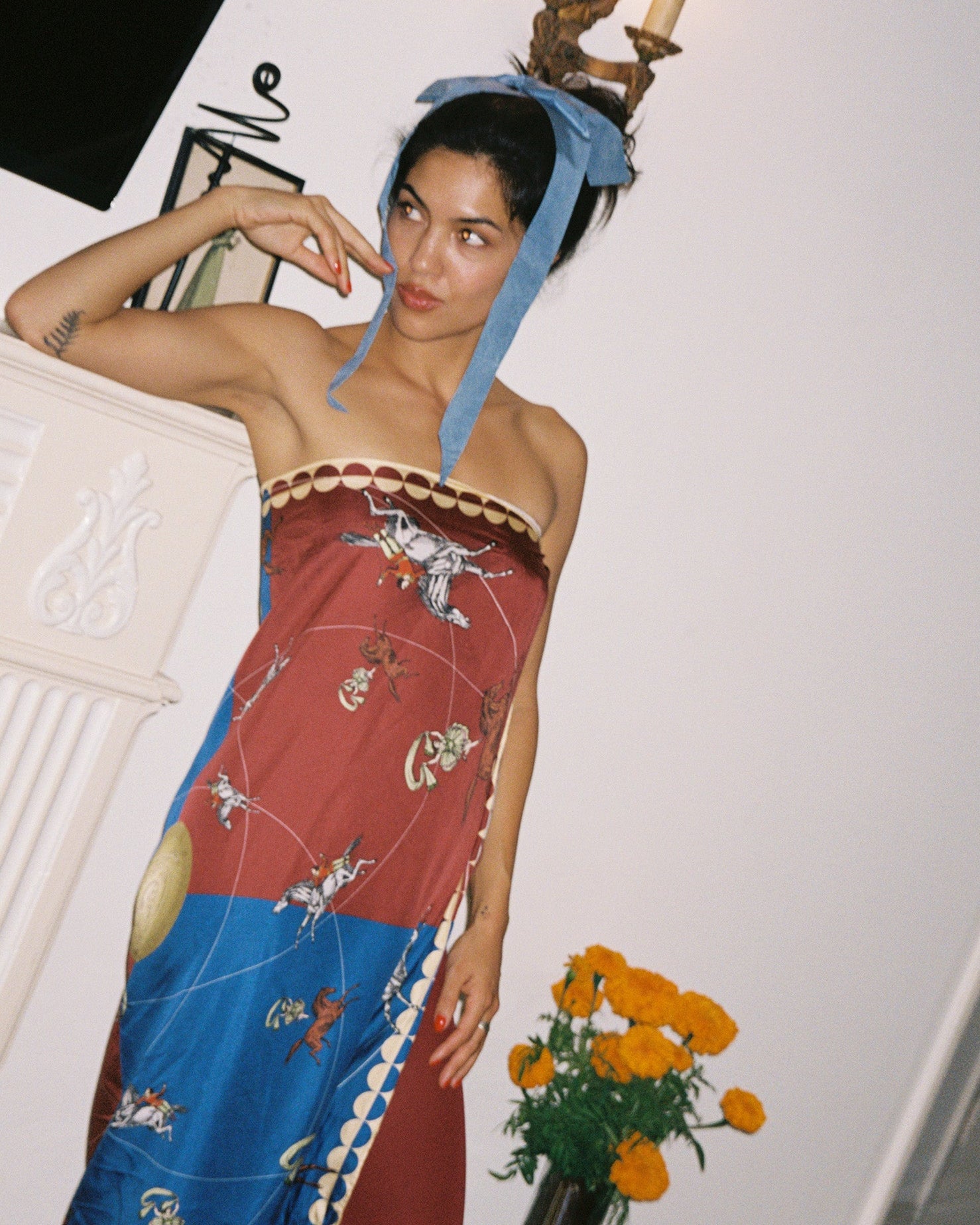 Woman wearing a colorful dress with a blue headscarf, standing next to a vase of flowers.