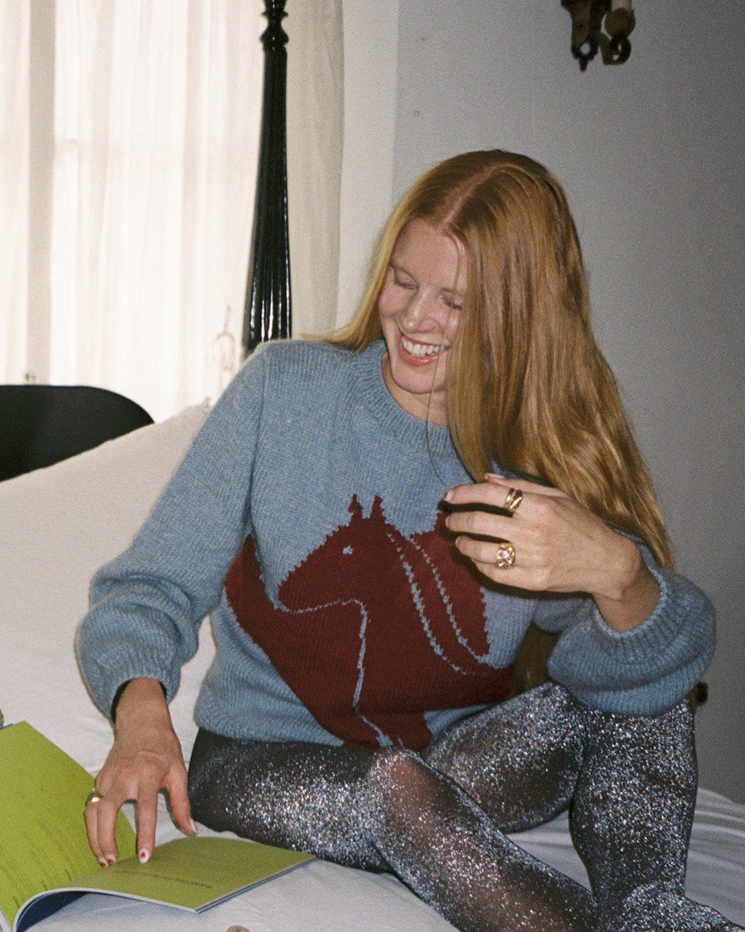 Woman in a blue sweater with a horse design, sitting on a bed reading a book.